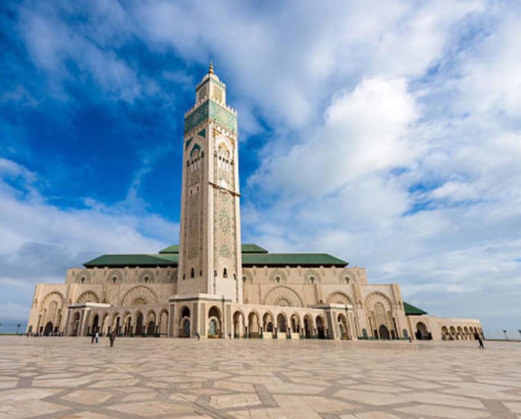 Fachada imponente da Mesquita Hassan II em Casablanca, Marrocos, destacando seu minarete alto sob um céu azul com nuvens. Em primeiro plano, o amplo pátio de mármore detalhado. Imagem promocional de roteiros culturais para o Marrocos pela KG Viagens.