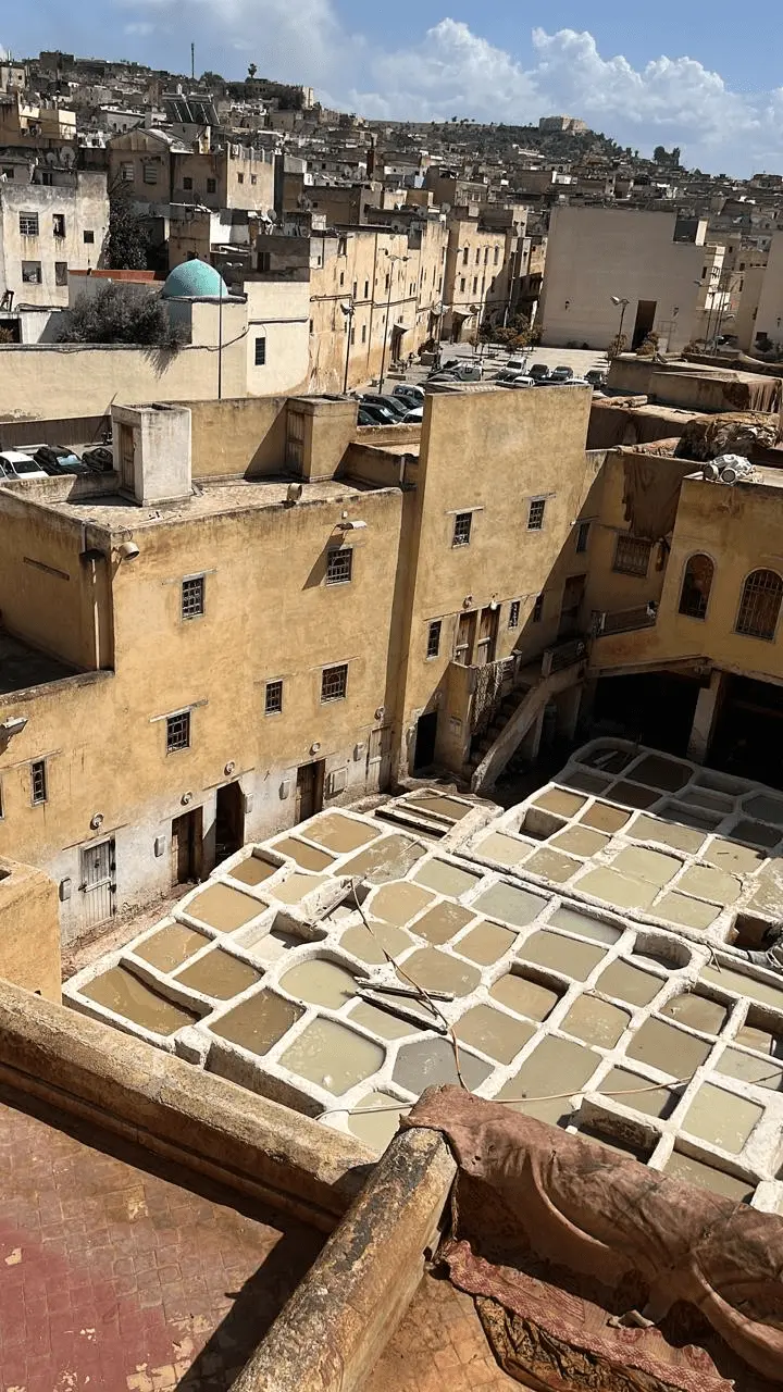 Vista panorâmica superior do Curtume Chouara na Medina de Fez, Marrocos. Vários tanques de pedra geométricos preenchidos com líquidos para o tingimento tradicional de couro aparecem cercados por edifícios antigos de arquitetura árabe. Destaque para os roteiros culturais exclusivos da KG Viagens.