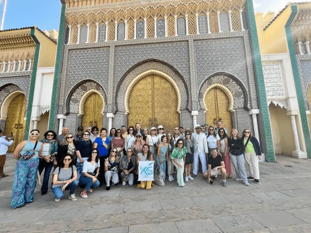 Grupo de turistas da agência KG Viagens reunido sorrindo e segurando a bandeira da empresa em frente aos grandes portões dourados de bronze do Palácio Real em Fez, Marrocos. Ao fundo, a fachada exibe mosaicos de azulejos coloridos e arquitetura geométrica islâmica.