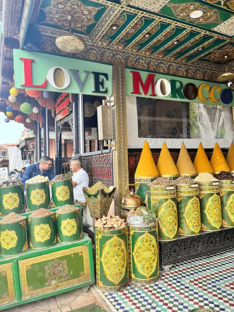 Loja tradicional de especiarias em um Souk de Marrakech, Marrocos, exibindo grandes cones de temperos coloridos e latas verdes ornamentadas com arabescos. No topo, um letreiro decorativo com a frase "LOVE MOROCCO" sob um teto trabalhado em madeira. Sugestão de compras e turismo cultural com a KG Viagens.