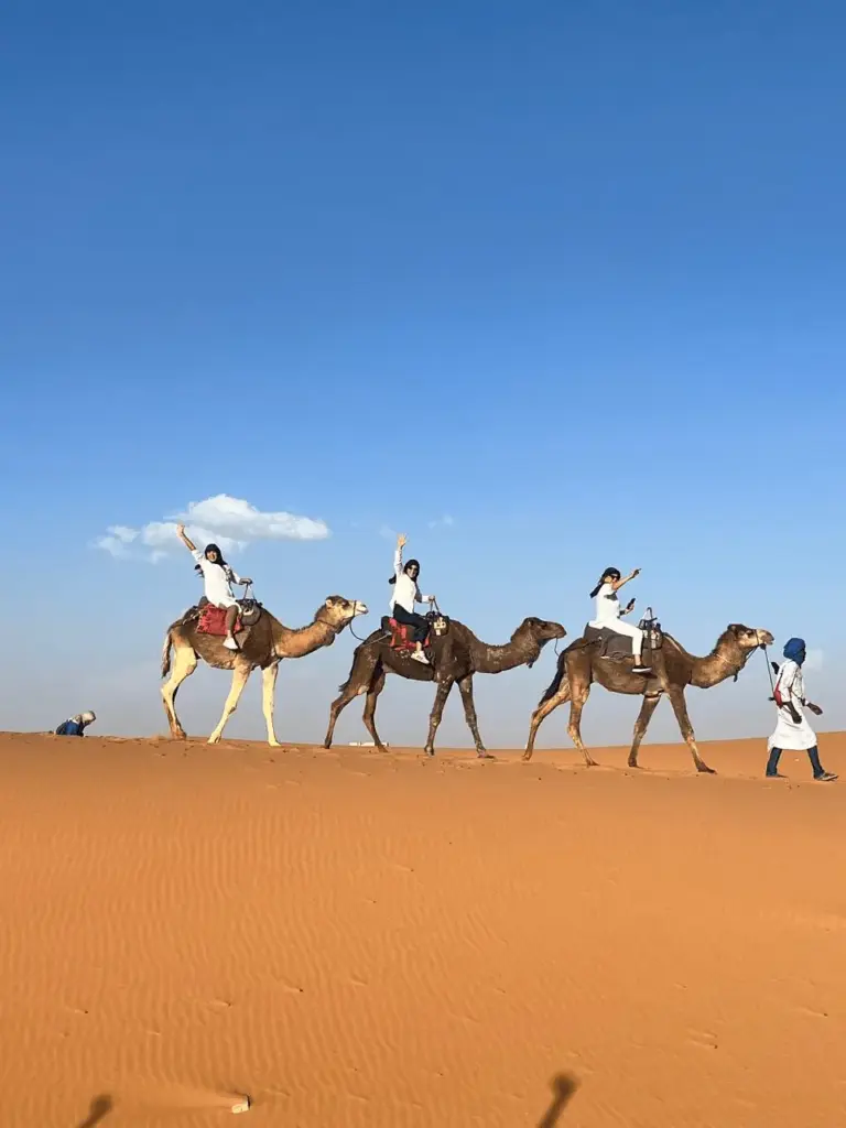 Três turistas da agência KG Viagens acenando felizes enquanto fazem um passeio de camelo pelas dunas alaranjadas do Deserto do Saara, em Merzouga, Marrocos. Um guia local com vestes tradicionais conduz a caravana sob um céu azul limpo e ensolarado.