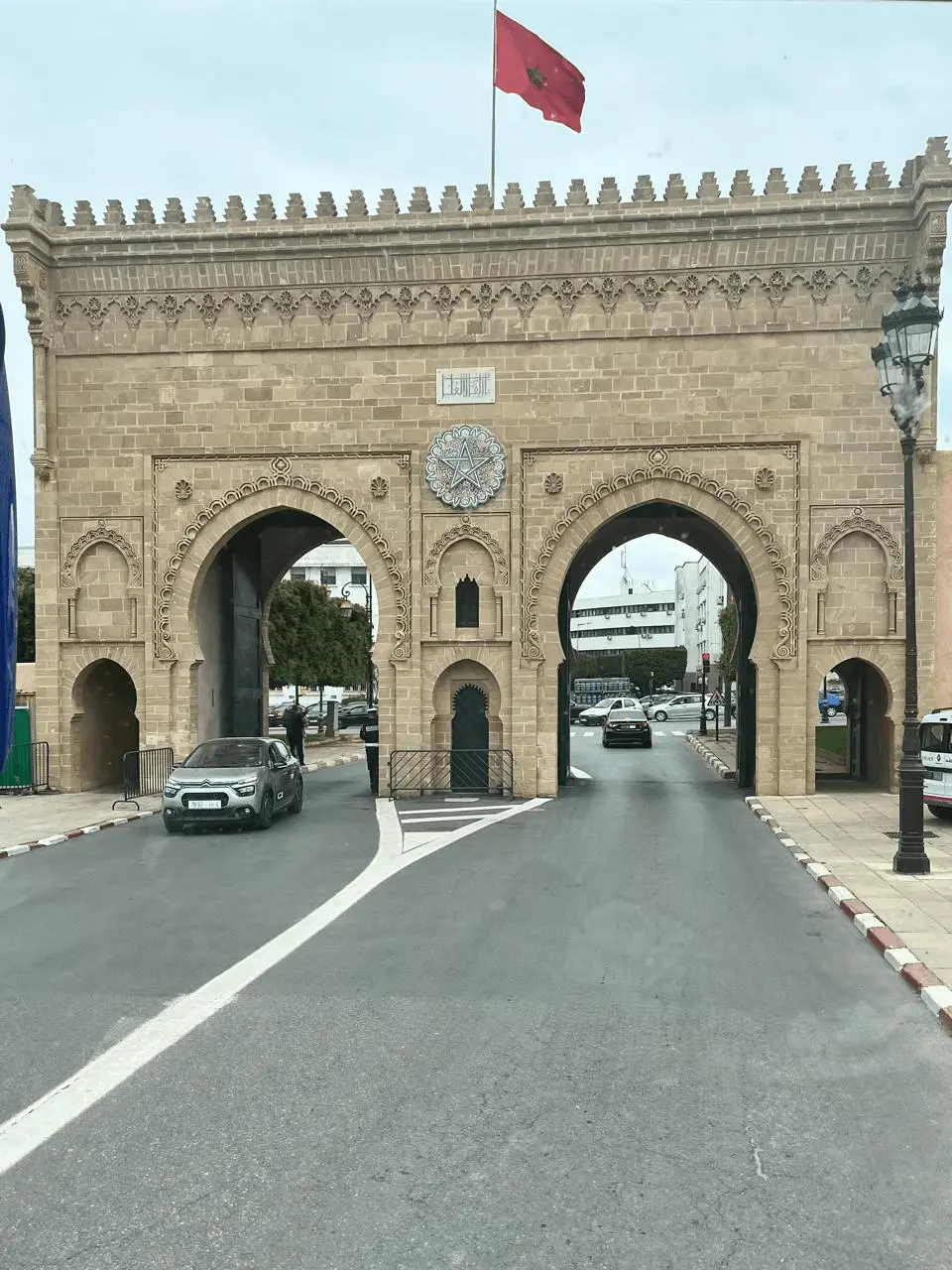 Portal monumental de pedra Bab er-Rouah em Rabat, Marrocos, com detalhes em arabescos, brasão da estrela verde e a bandeira marroquina hasteada no topo. Carros transitam sob os arcos da arquitetura islâmica tradicional. Sugestão de parada cultural em roteiros pelo Marrocos com a KG Viagens.